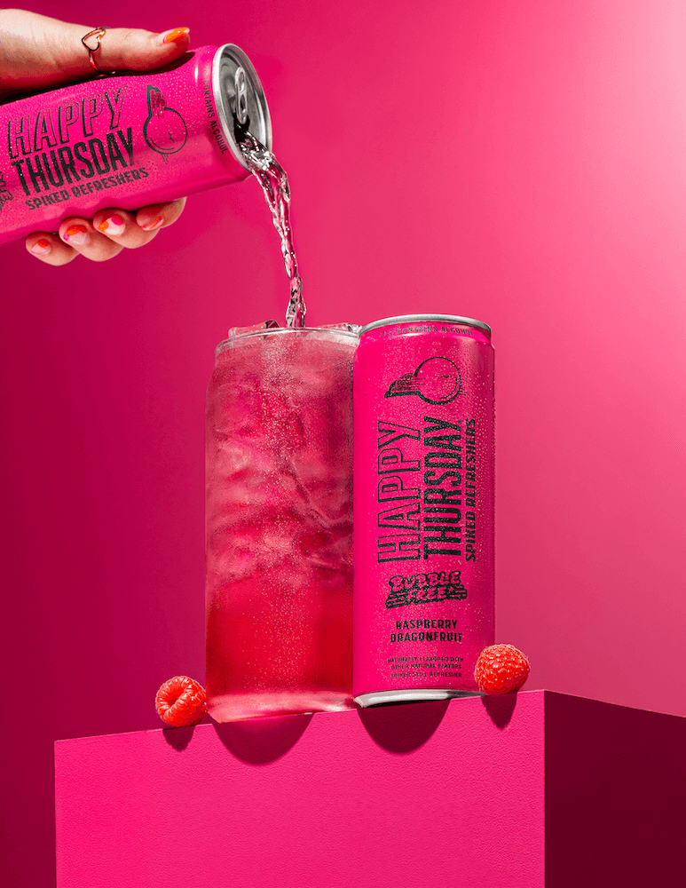 A can of Happy Thursday Raspberry dragonfruit Refresher is poured over ice into a glass, with Raspberry dragonfruit on a pink background.